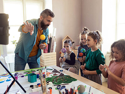 Ein Mann mit Bart und mehrere Kinder stehen um einen Tisch herum und basteln gemeinsam. Der Mann hält eine gelbe Kugel auf einem Stab, während die Kinder verschiedene Materialien und Werkzeuge in den Händen halten. Auf dem Tisch liegen bunte Farben, Pinsel, Papier und andere Bastelutensilien. Im Hintergrund sind weitere Bastelmaterialien und ein Fenster zu sehen.