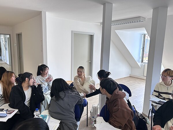 Mehrere Personen sitzen in einem hellen Raum auf Stühlen um einen Tisch. Sie unterhalten sich und blicken einander an. Auf dem Tisch liegen Hefte, Blätter und Schreibutensilien. Im Hintergrund sind Türen und große Fenster zu sehen, durch die Tageslicht einfällt. Die Wände und der Boden sind hell gehalten.