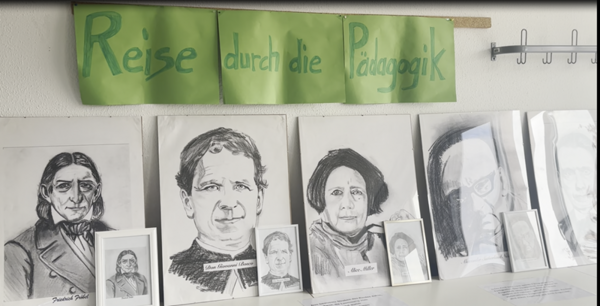 An einer Wand sind mehrere schwarz-weiße Porträtzeichnungen von historischen Persönlichkeiten der Pädagogik ausgestellt. Über den Zeichnungen hängt ein grünes Banner mit der handgeschriebenen Aufschrift 'Reise durch die Pädagogik'. Die Porträts sind in unterschiedlichen Größen gerahmt und nebeneinander auf einem Tisch oder Regal angeordnet. Die Szene vermittelt eine Ausstellung oder Präsentation im Rahmen des Tages der offenen Tür an der Euro Akademie Zwickau, die sich thematisch mit der Geschichte und Entwicklung der Pädagogik beschäftigt.