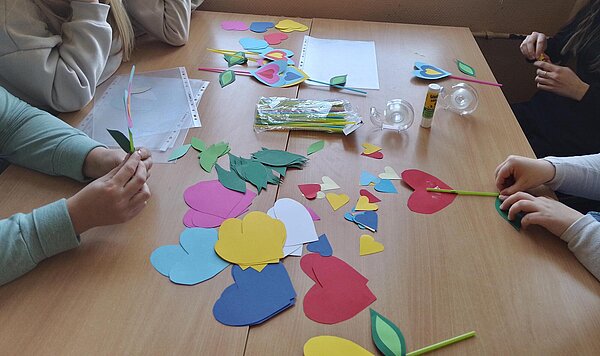 Mehrere Personen sitzen an einem Tisch und basteln mit buntem Papier. Es entstehen Blumen aus Herzformen, die auf Strohhalmen befestigt werden. Auf dem Tisch liegen Bastelmaterialien wie Klebestifte, Klebeband und Papierschablonen.