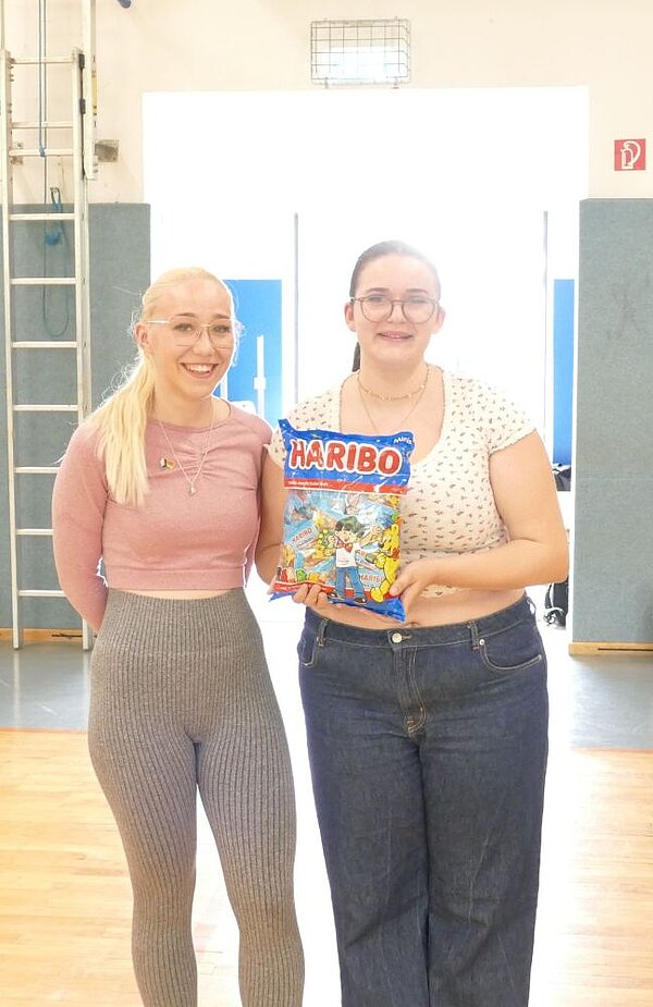 Zwei junge Frauen posieren lächelnd für ein Foto in einer Turnhalle. Die Frau links trägt ein rosafarbenes Oberteil und graue Leggings, während die Frau rechts ein weißes Oberteil mit Punkten und blaue Jeans trägt. Die Frau rechts hält eine Tüte mit der Aufschrift "HARIBO".