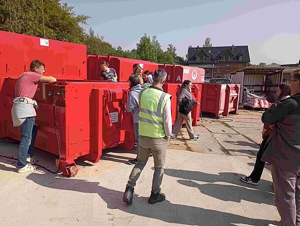 Schülerinnen und Schüler stehen vor großen roten Müllcontainern. 
