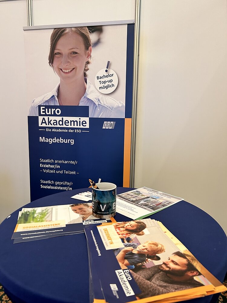 Im Vordergrund steht ein runder Tisch mit einem dunkelblauen Tischtuch. Darauf liegen mehrere Prospekte und Flyer, die das Logo und den Schriftzug »Euro Akademie« tragen. Eine Tasse mit Stiften steht ebenfalls auf dem Tisch. Im Hintergrund befindet sich ein aufgestelltes Roll-up-Banner mit dem Foto einer lächelnden jungen Frau im oberen Bereich. Darunter sind der Schriftzug »Euro Akademie – Die Akademie der ESO«, der Standort »Magdeburg« sowie Informationen zu angebotenen Ausbildungen und Studienmöglichkeiten in weißer Schrift auf blauem Grund zu sehen. Ein gelber Kreis hebt den Hinweis »Bachelor Top-up möglich« hervor.