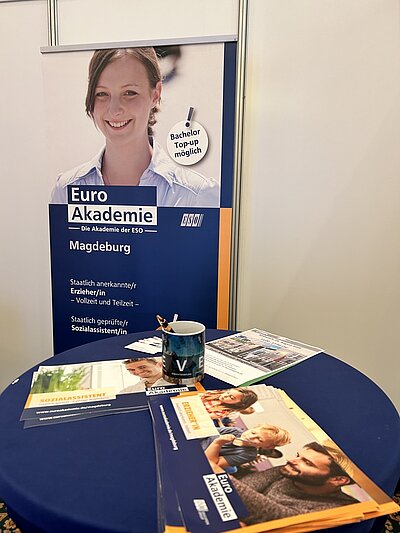 Im Vordergrund steht ein runder Tisch mit einem dunkelblauen Tischtuch. Darauf liegen mehrere Prospekte und Flyer, die das Logo und den Schriftzug »Euro Akademie« tragen. Eine Tasse mit Stiften steht ebenfalls auf dem Tisch. Im Hintergrund befindet sich ein aufgestelltes Roll-up-Banner mit dem Foto einer lächelnden jungen Frau im oberen Bereich. Darunter sind der Schriftzug »Euro Akademie – Die Akademie der ESO«, der Standort »Magdeburg« sowie Informationen zu angebotenen Ausbildungen und Studienmöglichkeiten in weißer Schrift auf blauem Grund zu sehen. Ein gelber Kreis hebt den Hinweis »Bachelor Top-up möglich« hervor.