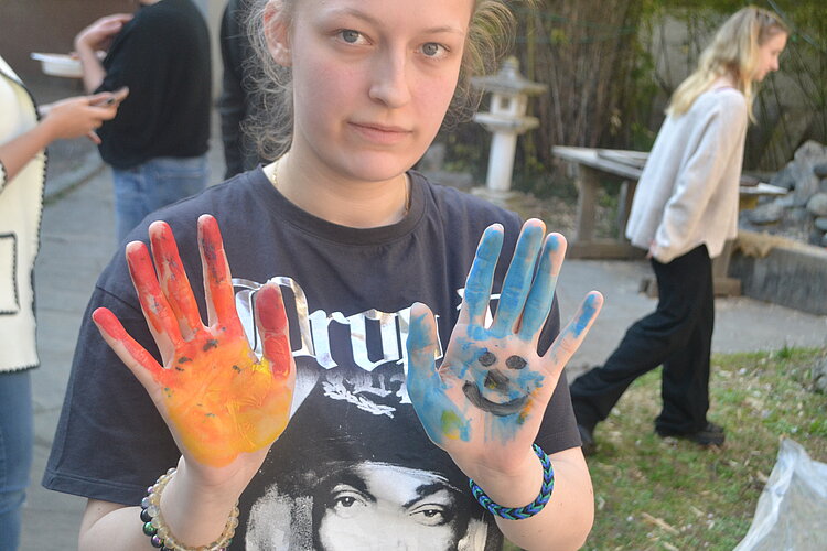 Malen als Selbsterfahrung mit Dorette Jansen Eine junge Person zeigt ihre Hände, die bunt mit Farbe bemalt sind. Auf der linken Hand befinden sich warme Farbtöne wie Rot, Orange und Gelb, während die rechte Hand blau bemalt ist und ein lächelndes Gesicht zeigt. Im Hintergrund sind weitere Personen sowie ein Garten zu erkennen.