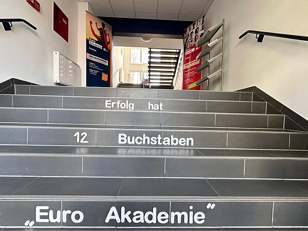 Eine grau geflieste Treppe im Eingangsbereich der Euro Akademie, auf deren Stufen steht: "Erfolg hat 12 Buchstaben: Euro Akademie".