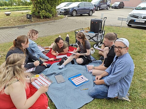 Sommerliches Abschlussfest an der Euro Akademie Jena Schülerinnen und Schüler sitzen auf einer Decke auf der Wiese, alle haben Blätter und Stifte in der Hand und spielen gemeinsam.