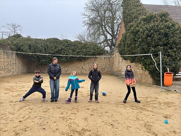 Winter-Sprachferien 2025: „Living in Fairy Tales“ Fünf Kinder unterschiedlichen Alters in Winterjacken stehen auf einem sandigen Volleyballfeld. Ein Kind trägt einen großen Stein. Auf dem Boden liegt ein blauer Ball. Am Spielfeldrand ist ein orangefarbener Mülleimer und im Hintergrund eine von üppigen Büschen bewachsene Mauer zu sehen.