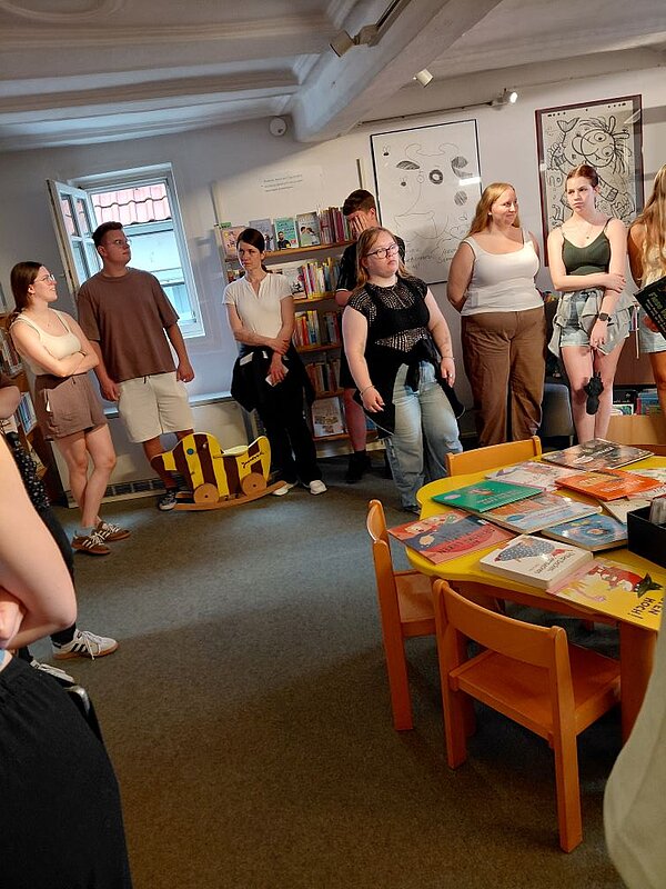 Eine Gruppe von jungen Erwachsenen steht eng beieinander in einem Raum mit Kinderbüchern, Spielzeug und bunten Möbeln. Im Hintergrund sind Regale mit Büchern und ein Fenster zu sehen.