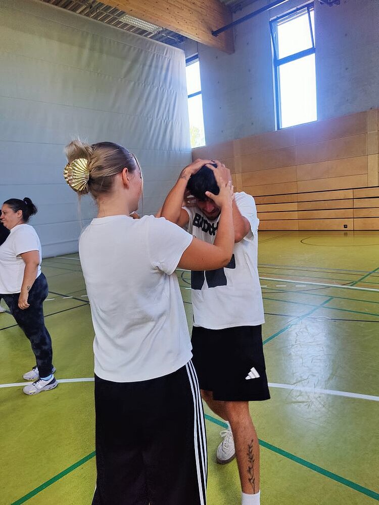 Zwei Personen in einer hellen Sporthalle mit Holzpaneelen an den Wänden. Eine Frau mit einer goldenen Haarklammer, weißem T-Shirt und schwarzer Hose ergreift die Arme eines Mannes mit schwarzer Shorts und weißem T-Shirt, der sich diese schützend vor das Gesicht hält. Im Hintergrund ist eine weitere Person zu sehen.