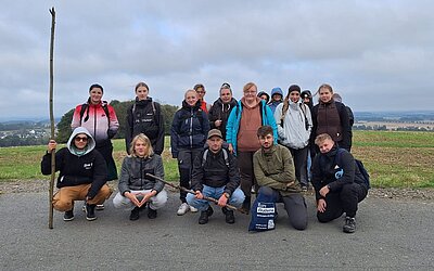 Eine Gruppe junger Erwachsener und Auszubildender steht gemeinsam draußen auf einer Straße vor einer weiten Feldlandschaft. Sie tragen Rucksäcke und wetterfeste Kleidung, einige knien vorne, andere stehen hinten.