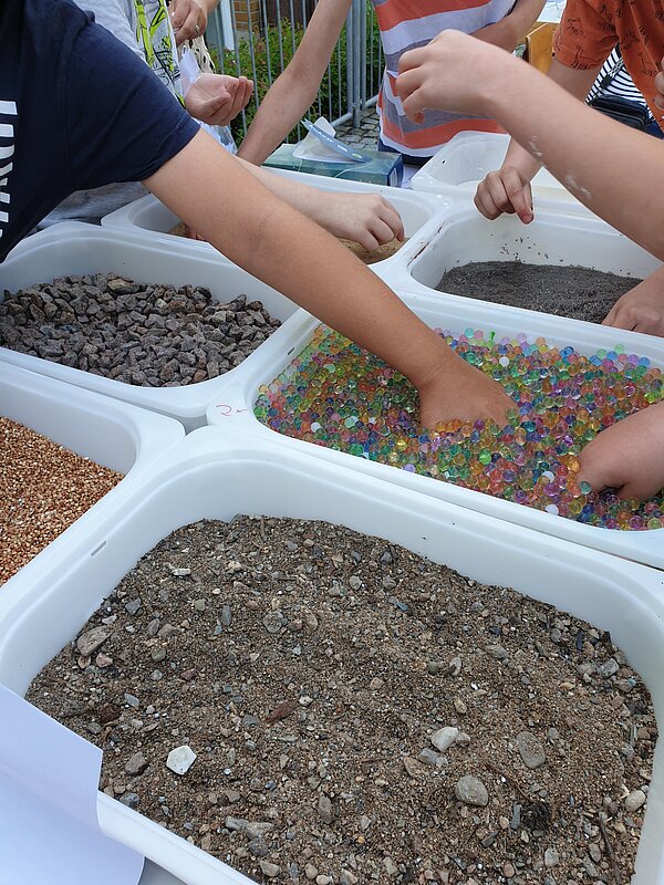 Ein erlebnisreicher Vormittag voller Spiel, Kreativität und Gemeinschaft Kinderhände greifen in verschiedene Fühlwannen mit Steinen, bunten Wasserperlen und Sand an einem Mitmachstand.
