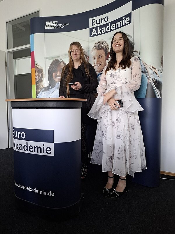 Zwei Absolventinnen, eine in schönem weißen Kleid, stehen am Pult.