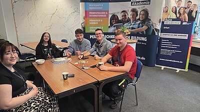 Mehrere Auszubildende der Euro Akademie Erfurt sitzen um zwei zusammengeschobene Holztische in einem hellen Raum. Auf den Tischen liegen verschiedene Gegenstände, darunter bunte Stifte, Papier und ein Becher. Im Hintergrund stehen zwei große Roll-up-Banner mit dem Schriftzug 'Euro Akademie' und weiteren Texten. Die Wände sind hell und mit einer abstrakten, bunten Linienzeichnung verziert. Die Azubis tragen Namensschilder und posieren für ein Gruppenbild.