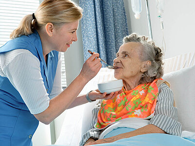 Eine Pflegekraft füttert eine ältere Patientin im Krankenhausbett mit einem Löffel aus einer Schüssel.