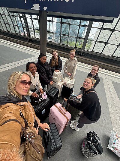 Sieben junge Frauen stehen mit ihren Koffern auf einem Bahnsteig und lächeln in die Kamera.