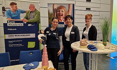 Drei Personen stehen vor Roll-ups der Euro Akademie; eine Person hat eine Puppe im Arm.
