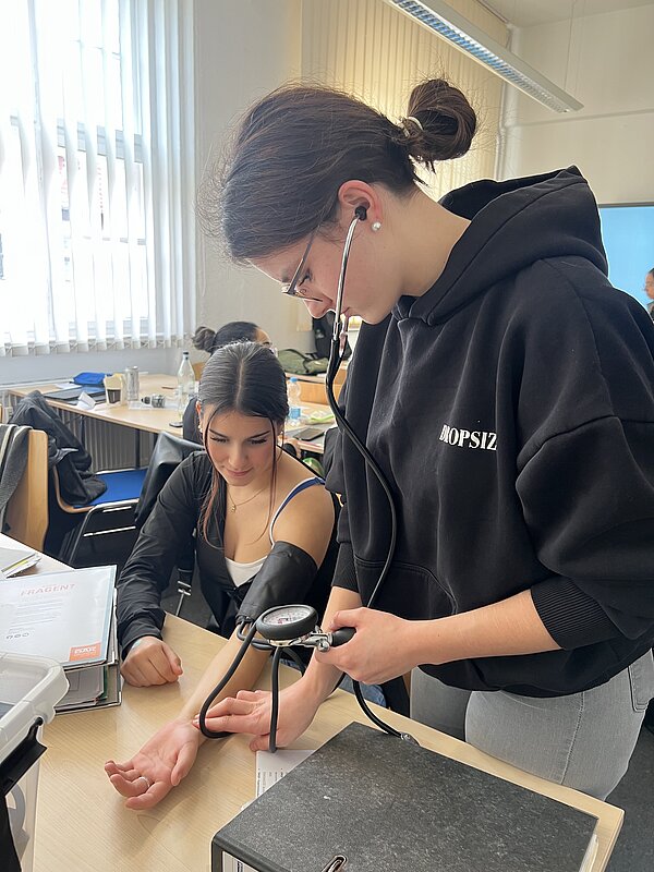 In einem Unterrichtsraum misst eine Schülerin mit Stethoskop und Blutdruckmanschette den Blutdruck einer Mitschülerin, die mit freiem Arm am Tisch sitzt.