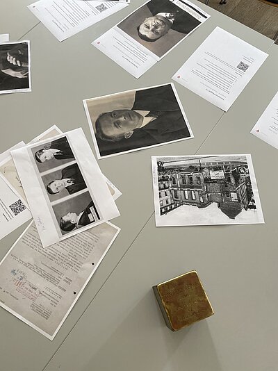 Auf einem Tisch liegen historische Porträtfotografien, Dokumente, Arbeitsblätter mit QR-Codes und das Schwarz-Weiß-Foto eines Gebäudes. Im Vordergrund befindet sich ein goldfarbener Erinnerungsstein mit einer Inschrift für Toni Rosenbaum.
