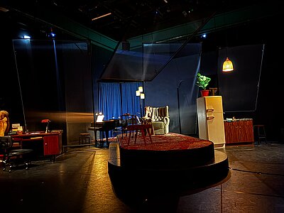 Eine schwach beleuchtete Theaterbühne. In der Mitte ein Sessel auf einem erhöhten Podest, dahinter ein Klavier vor einem blauen Vorhang. Links ein altmodischer Schreibtisch. Rechts ein Kühlschrank und eine Küchenzeile.