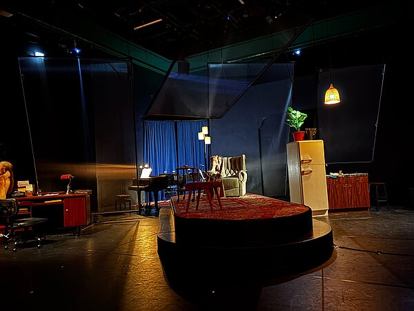 Eine schwach beleuchtete Theaterbühne. In der Mitte ein Sessel auf einem erhöhten Podest, dahinter ein Klavier vor einem blauen Vorhang. Links ein altmodischer Schreibtisch. Rechts ein Kühlschrank und eine Küchenzeile.