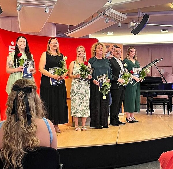 Feierlicher Abschied: Exmatrikulation der Absolvent*innen 2025 Eine Gruppe von Personen steht auf einer Bühne und hält Blumen und Zertifikate. Sie sind elegant gekleidet und lächeln. Im Hintergrund ist ein rotes Banner und ein Klavier zu sehen.