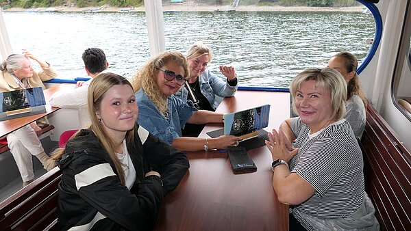 Mehrere Frauen sitzen gemeinsam an einem Tisch an Bord eines Ausflugsschiffes und lächeln in die Kamera, während im Hintergrund der Stausee zu sehen ist.