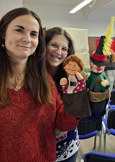 Zwei lächelnde junge Frauen halten bunte Handpuppen in einer festlich geschmückten Umgebung.