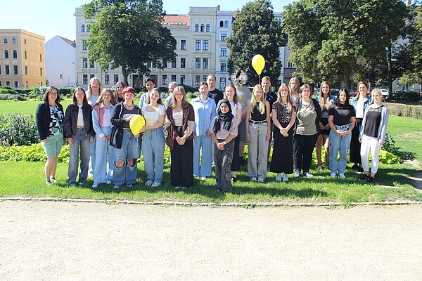 Herzlich Willkommen zum ersten Schuljahr an der Euro Akademie Görlitz! Auf einer sonnigen Wiese inmitten der Stadt haben sich die neuen Auszubildenden für ein Gruppenfoto versammelt. Zwei gelbe Luftballons haben es auch auf das Bild geschafft.