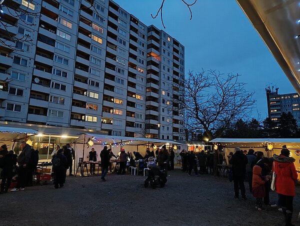 Abendliche Szene eines kleinen Weihnachtsmarkts im Innenhof hoher Wohnblocks, mehrere festlich beleuchtete Stände mit vielen Besuchenden, einige mit Kinderwagen und in Winterkleidung.