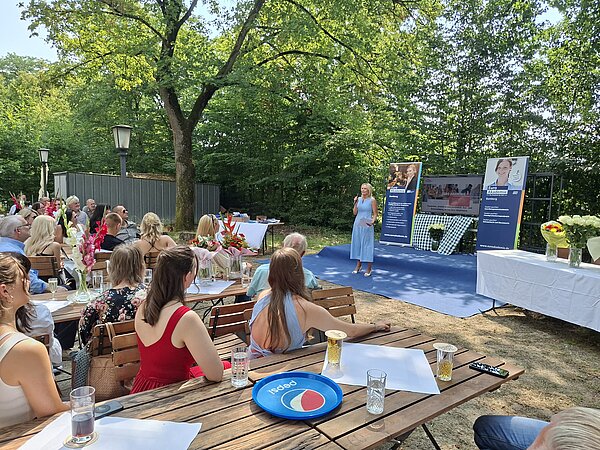 Eine Person spricht auf einer Bühne bei einer Abschlussveranstaltung im Freien. Die Gäste sitzen an Tischen vor der Bühne. Blumensträuße, Roll-up-Banner und eine festlich gestaltete Umgebung unter Bäumen prägen die Szene.