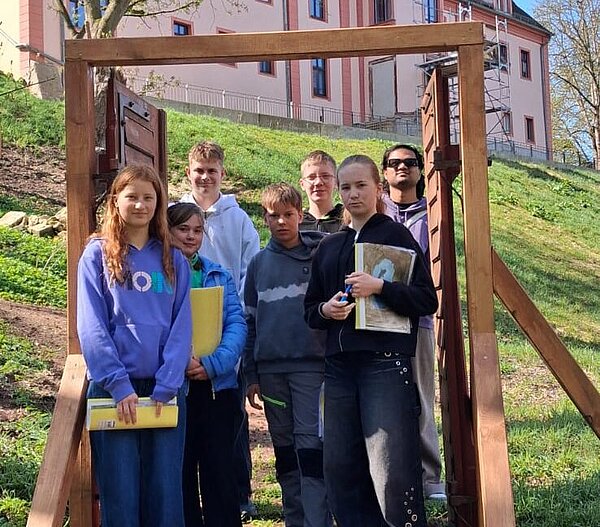 Gruppe von Jugendlichen steht im Freien unter einem hölzernen Rahmen vor einem historischen rosa Gebäude am Hang.