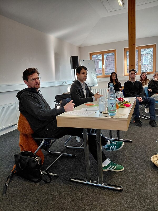 Eine Gruppe von Auszubildenden sitzt in einem Besprechungsraum. Im Vordergrund sitzen zwei Männer an einem Tisch, einer trägt einen Kapuzenpullover, der andere einen Anzug. Auf dem Tisch liegen Papiere und eine Wasserflasche. Im Hintergrund sitzen ihnen mehrere Personen gegenüber; eine Tafel und Fenster sind zu sehen. Es scheint sich um eine Diskussions- oder Präsentationssituation zu handeln.