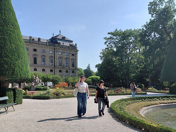 Zwei Personen spazieren durch den barocken Hofgarten der Würzburger Residenz. Im Hintergrund erhebt sich das imposante Gebäude der Residenz mit seinen symmetrischen Fenstern und dem Mansarddach. Der Garten ist mit gepflegten Hecken, Blumenbeeten und hohen Bäumen gestaltet.