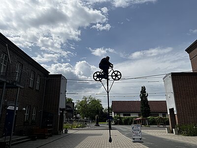 Jemand fährt auf einem Fahrrad über ein Seil, das mehrere Meter über dem Boden gespannt ist; der Himmel dahinter ist blau und es sind weiße Wolken zu sehen.