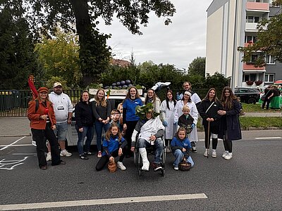 Gruppenbild mit lächelnden Menschen auf einer Straße. Mehrere Personen sind mit weißen Kitteln verkleidet und eine Person sitzt mit Bandagen im Rollstuhl. Hinter der Menschengruppe steht ein Auto mit selbstgebastelten Weintrauben auf dem Dach. Im Hintergrund sind Bäume und Häuser zu sehen.