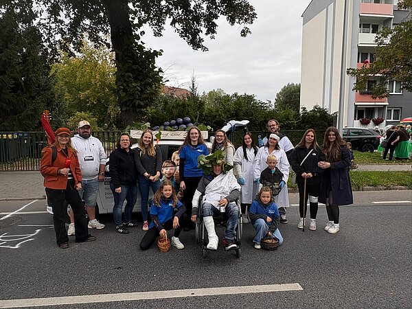Gruppenbild mit lächelnden Menschen auf einer Straße. Mehrere Personen sind mit weißen Kitteln verkleidet und eine Person sitzt mit Bandagen im Rollstuhl. Hinter der Menschengruppe steht ein Auto mit selbstgebastelten Weintrauben auf dem Dach. Im Hintergrund sind Bäume und Häuser zu sehen.