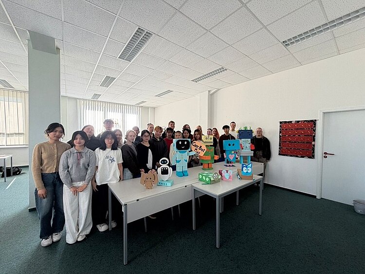 Eine Gruppe von Auszubildenden der Euro Akademie Bitterfeld-Wolfen steht in einem hellen Raum mit weißen Wänden und Decke. Im Vordergrund stehen auf zusammengeschobenen Tischen mehrere bunte, selbstgebastelte Roboterfiguren aus Karton und Papier. Die Figuren haben verschiedene Formen, Farben und aufgemalte Gesichter. Im Hintergrund sind große Fenster mit senkrechten Lamellen und eine geschlossene weiße Tür zu sehen. An der Wand hängt ein rechteckiges, rotes Objekt mit schwarzen Elementen.