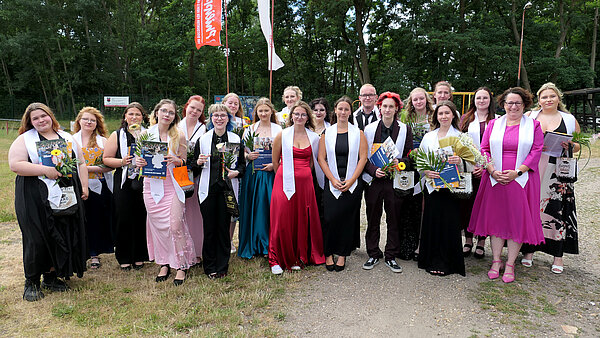 Auf einem grasbewachsenen Kiesweg vor Bäumen steht eine Gruppe junger Menschen in festlicher Kleidung. Sie tragen weiße Schärpen um die Schultern. Einige Menschen halten Blumen und Zeugnismappen in den Händen.