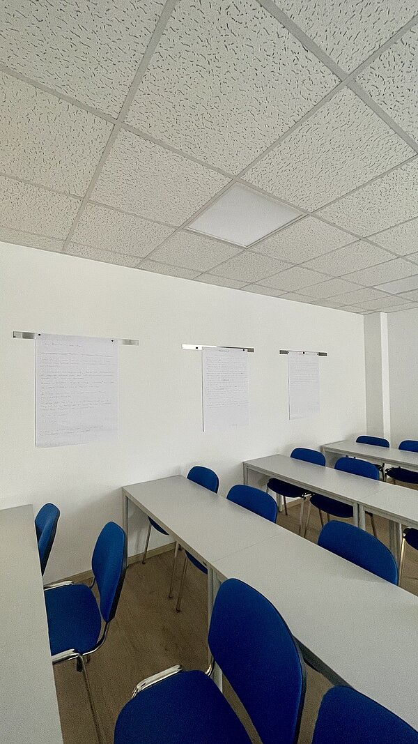 Heller Unterrichtsraum mit weißen Tischen, blauen Stühlen und mehreren beschriebenen Papierbögen an der Wand.