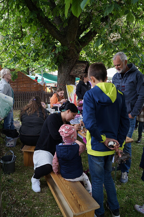Personen sitzen und stehen um einen Tisch unter einem Baum herum, an dem offenbar Kinderschminken angeboten wird. Erwachsene schminken Kinder, während weitere Personen im Hintergrund stehen. Die Atmosphäre ist entspannt und gesellig auf einer Grünfläche im Freien.