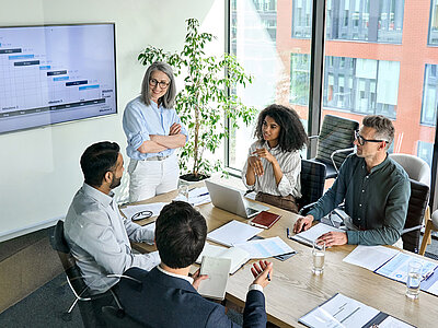 Eine Gruppe von fünf Personen sitzt um einen Konferenztisch in einem modernen Besprechungsraum. Eine Frau steht und präsentiert etwas, während die anderen vier Personen zuhören und Notizen machen. Auf dem Tisch liegen verschiedene Dokumente, Laptops und Gläser mit Wasser. Im Hintergrund hängt ein großer Bildschirm mit einem Projektplan oder Gantt-Diagramm. Eine große Pflanze steht neben dem Bildschirm.