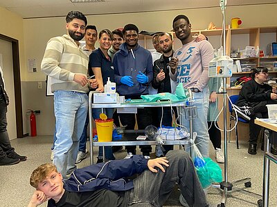 Gruppenbild eines medizinischen Workshops: Die Teilnehmer:innen stehen lachend um einen Arbeitstisch mit medizinischen Materialien. Einige tragen Handschuhe, andere halten medizinisches Werkzeug in der Hand. Einer der Teilnehmer liegt entspannt vorne auf dem Boden und posiert für das Foto.
