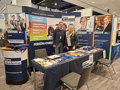 Messestand der Euro Akademie mit Informationsmaterialien, Werbebannern und zwei Mitarbeitenden vor Ort.