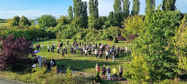 Wikinger-Sportfest: Pflegeazubis erobern die nordische Arena Zahlreiche Menschen stehen und bewegen sich auf einer großen grünen Wiese, umgeben von Bäumen. Es wirkt wie eine gemeinsame Sport- oder Bewegungsaktion im Freien.