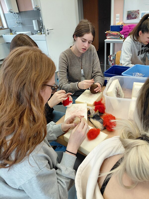 Jugendliche sitzen dicht beisammen an einem Tisch und formen mit Filznadeln und Wolle kleine Dekoobjekte, vor ihnen liegen rote und weiße Wollstücke.