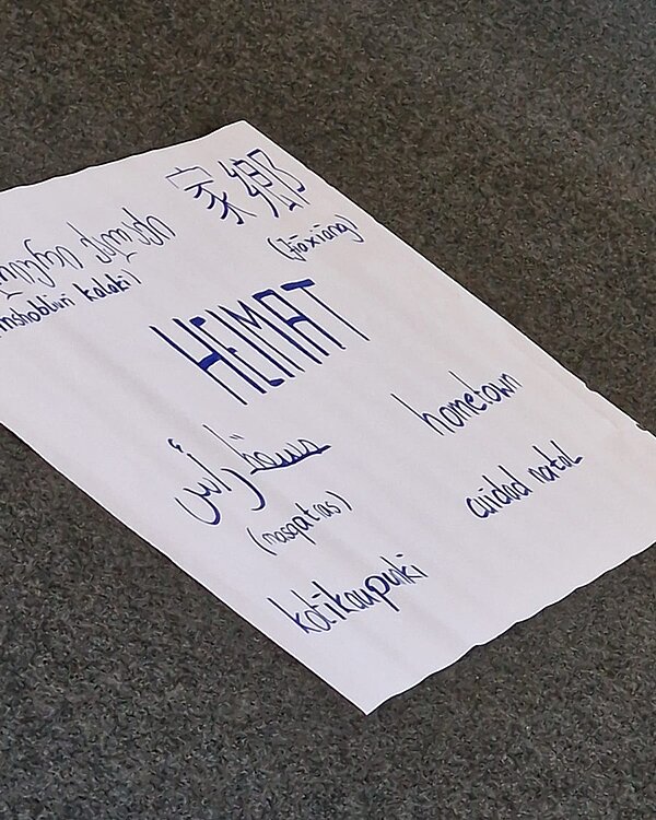Theaterbesuch und Workshop Großes Blatt, auf dem mittig das Wort Heimat auf Deutsch steht und rundherum in weiteren Sprachen.