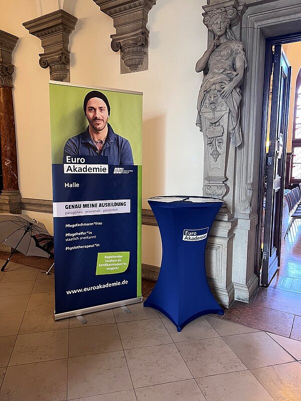In einem Steinkorridor mit Säulen und Statuen an der Wand stehen ein Roll-up und ein blau bespannter Stehtisch der Euro Akademie Halle.