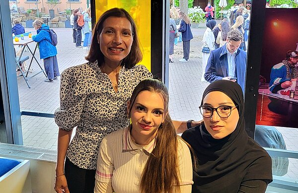 Spanischunterricht mit Astor Piazzolla: Yo soy María! Die Lehrerin hat sich neben zwei sitzenden Schülerinnen, von denen eine ein Kopftuch trägt, für ein Foto positioniert. Hinter der Gruppe sieht man verschiedene Zuschauer*innen durch ein Fenster im Innenhof der Oper in Gruppen und an Tischen stehen.