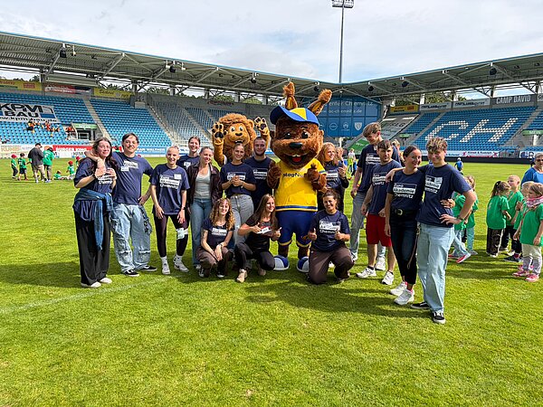 Gruppenfoto von Mitarbeitenden und Teilnehmenden der Euro Akademie Chemnitz auf dem Stadionrasen gemeinsam mit zwei großen Maskottchen.